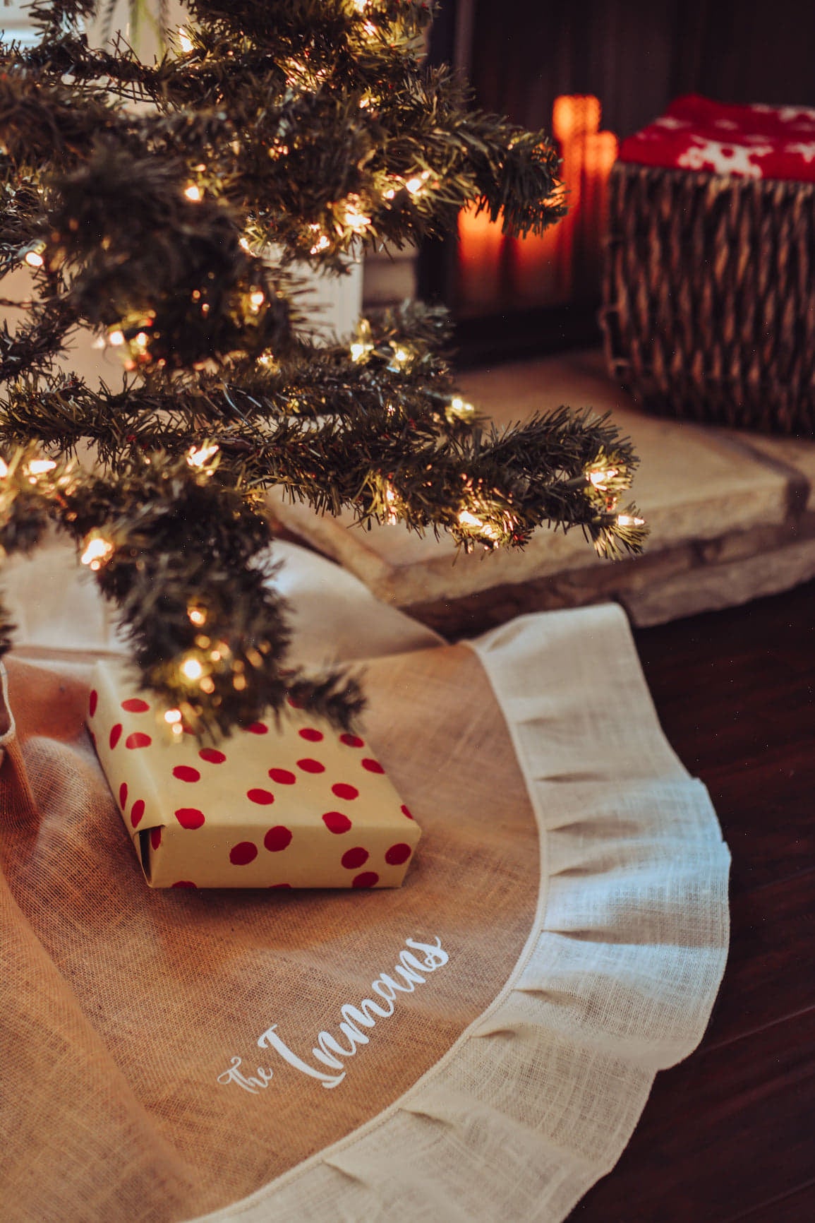 Tan Burlap Christmas Tree Skirt with White outer ruffle. Skirt is personalized with family name, The Inmans.  Tree Skirt is accented under a lit tree with gift.