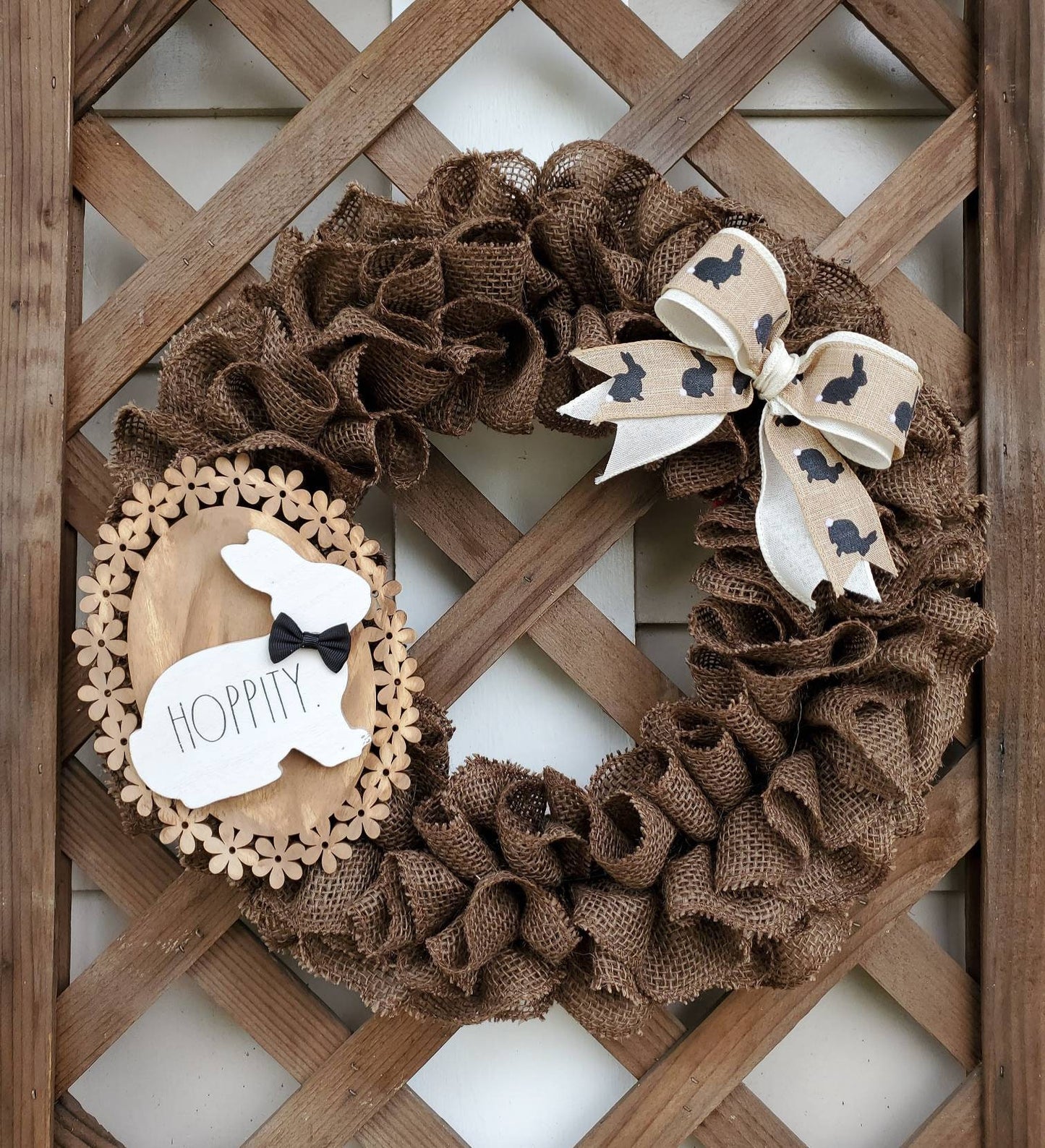 Brown Burlap Easter Wreath with cream & tan bow adorned with brown bunny silhouettes. Made complete with wood flower base sign accented with white rabbit with black bow tie. Set upon lattice wall..