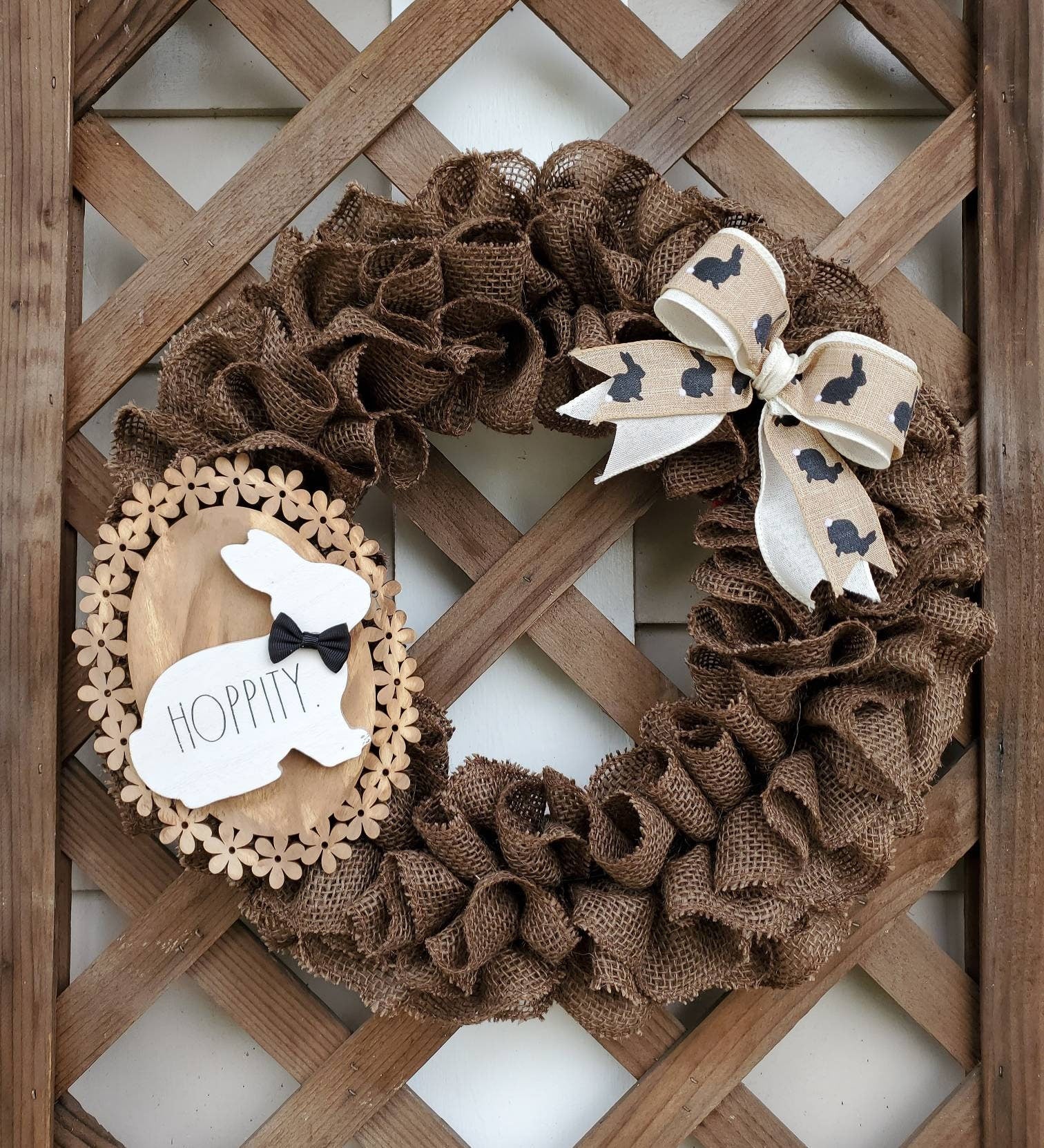 Brown Burlap Easter Wreath with cream & tan bow adorned with brown bunny silhouettes. Made complete with wood flower base sign accented with white rabbit with black bow tie. Set upon lattice wall..