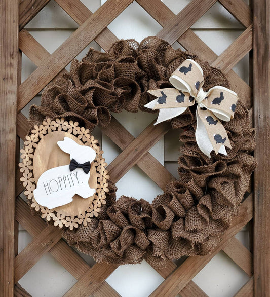 Brown Burlap Easter Wreath with cream & tan bow adorned with brown bunny silhouettes. Made complete with wood flower base sign accented with white rabbit with black bow tie. Set upon lattice wall..