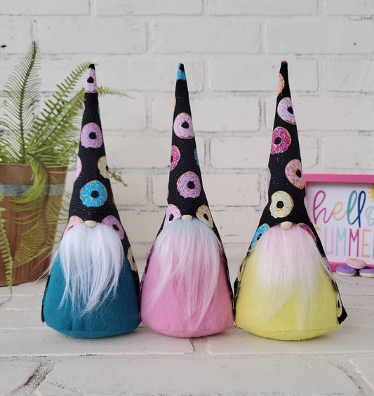 Donut Gnome collection. Adorned with black base hats accented with variety colored donuts in pinks, yellows and blues. First with white beard and teal body, second with ombre beard and pink body and lastly pink beard with yellow body. These handmade KyElle Kreations gnomes measure 9 inches.. Displayed with faux foliage and seasonal accent decor.
