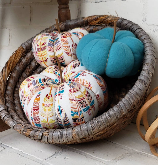 Pumpkin trio set. One large and one small crafted from a cream-colored base with bold colorful feathers in burgundy, teal and gold hues. Third pumpkin in solid teal. Displayed in hand woven basket