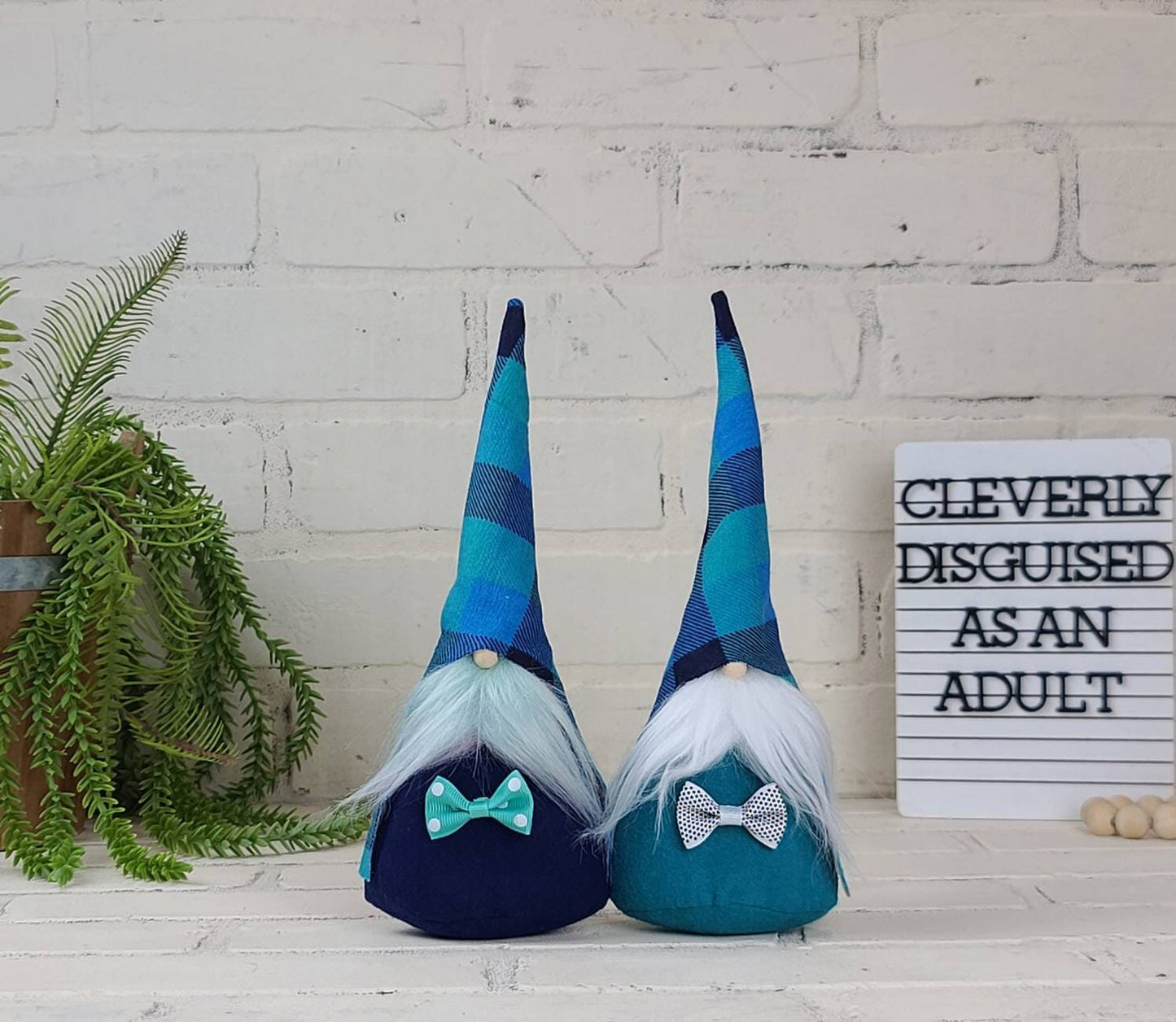 Blue Buffalo Plaid Gnomes. Plaid with navy and teal hue plaid hats, one with lite blue beard and navy body. Other with white beard and teal body. These handmade KyElle Kreations gnomes measures 9 inches.. Displayed with faux foliage and seasonal accent decor. 
