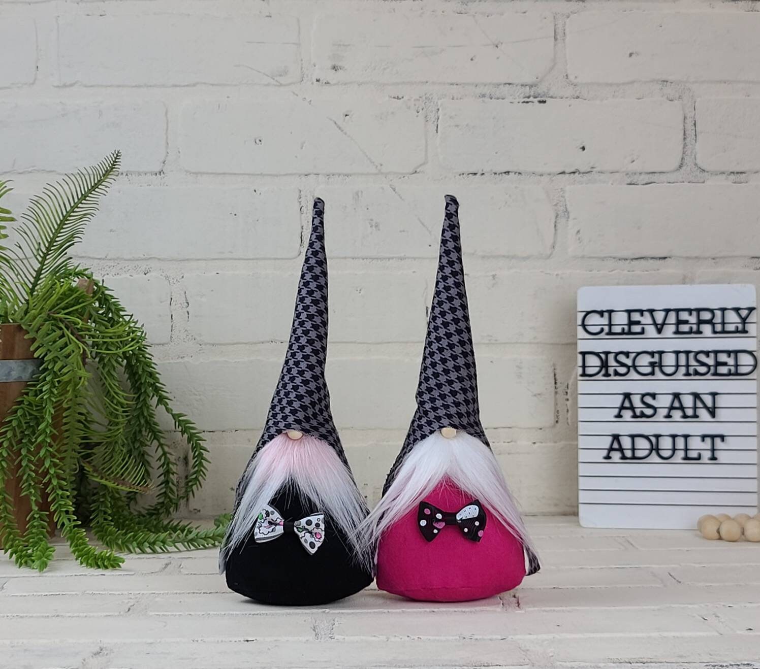 Checkered Gnomes with Black & Gray checkered hats. One with light pink beard and back body. Other with white beard and pink body, both adorned with bow ties. This handmade KyElle Kreations gnome measures 9 inches.. Displayed with faux foliage and seasonal accent decor. 