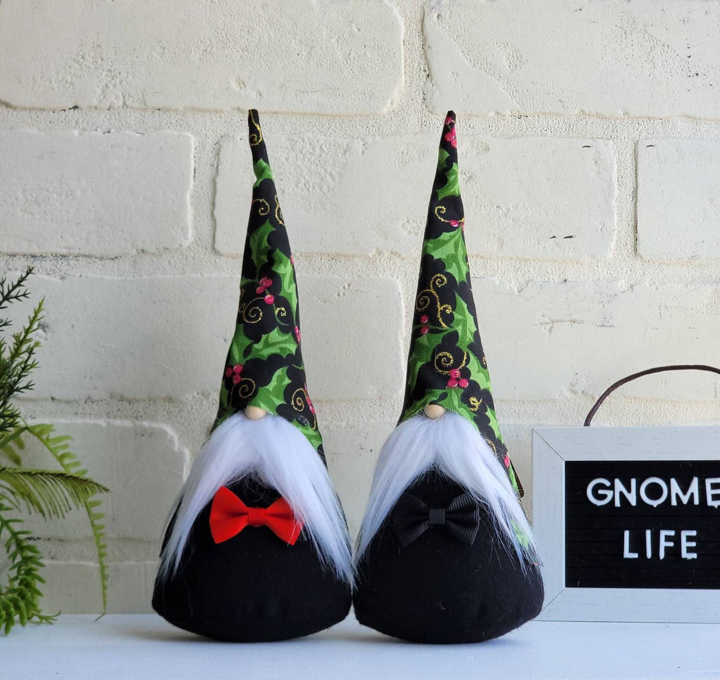Christmas Holly Gnomes with white beards & black bodies. One with red bow tie and the other with black. Thiese handmade KyElle Kreations gnomes measure 9 inches.. Displayed with faux foliage and seasonal accent decor. 